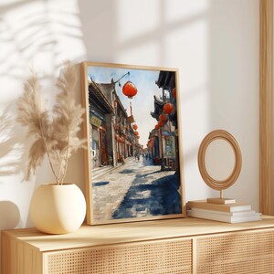 May include: Framed watercolor print of a traditional Asian street scene. The artwork features a cobblestone street lined with buildings and red lanterns. The print is displayed on a wooden cabinet with a vase and a decorative mirror.