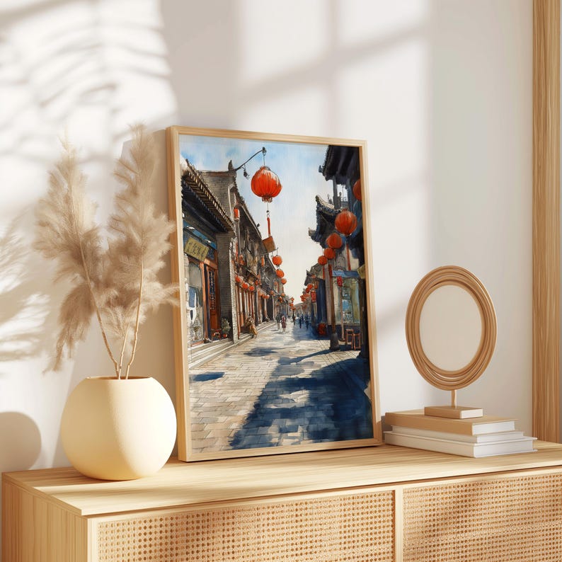 May include: Framed watercolor print of a traditional Asian street scene. The artwork features a cobblestone street lined with buildings and red lanterns. The print is displayed on a wooden cabinet with a vase and a decorative mirror.