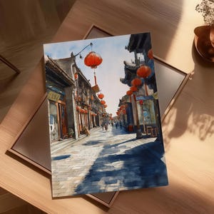 May include: Watercolor painting of a street scene in a traditional Asian town. The artwork depicts buildings with ornate roofs, red lanterns, and a cobblestone street. The painting is set in a framed canvas, resting on a wooden table.
