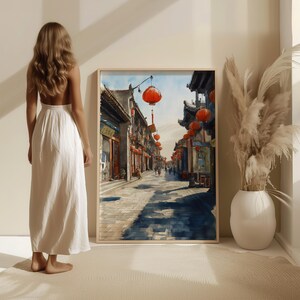 May include: A framed watercolor painting depicts a traditional Asian street scene with red lanterns. The artwork is displayed in a light-colored wooden frame. A woman in a white dress stands nearby, and a vase with decorative plants is in the corner.