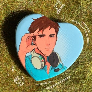 May include: Heart-shaped button with a cartoon illustration of a person wearing a headset. The person has brown hair and is wearing a blue shirt. The background is light blue with a halftone pattern. White doodles surround the button.