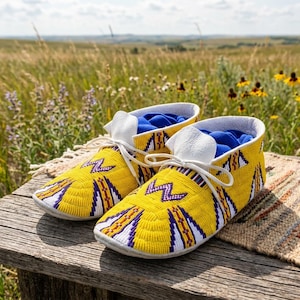 May include: A pair of yellow moccasins featuring intricate beadwork in purple, white, and orange geometric patterns. The footwear includes white leather laces and a bright blue interior lining, showcasing a detailed hand-beaded design on the exterior.