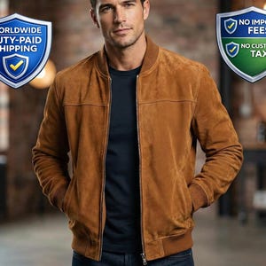 May include: A brown suede bomber jacket with a ribbed collar, cuffs, and hem. The jacket has a zipper closure and two front pockets. The model is wearing a navy blue t-shirt and blue jeans. The background is a brick wall.