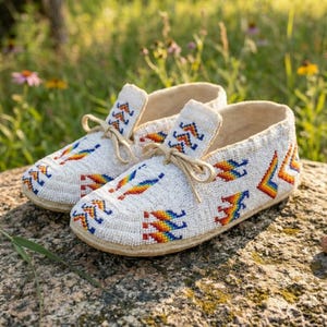 May include: A pair of white moccasins featuring intricate beadwork in a vibrant rainbow-colored geometric pattern. Each shoe includes a matching white leather lace tied in a bow at the front, with a light-colored sole and a soft interior.