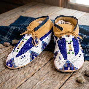 Op de afbeelding: Een paar mocassins van beige suède met ingewikkeld kralenwerk in blauwe, witte, groene en gele geometrische patronen. Het schoeisel is voorzien van een donkerblauwe rand rond de kraag en dunne beige suède veters gestrikt aan de voorzijde.