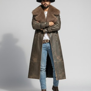 May include: A long, brown leather coat with a fur collar, worn with a black hat, blue jeans, and brown boots. The coat has a double-breasted design and a belt at the waist. The model is standing with arms crossed.