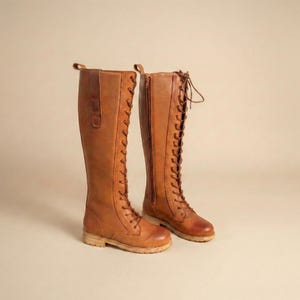 Peut inclure: Une paire de longues bottes en cuir marron, arrivant au genou. Les bottes ont un la&ccedil;age, une fermeture &eacute;clair lat&eacute;rale et une petite languette en haut. Les semelles sont de couleur beige clair.