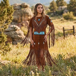 May include: A brown suede dress with fringe detailing and turquoise and white bead accents. The dress features a round neckline and a fitted waist. The model is standing in a field with a mountain backdrop.