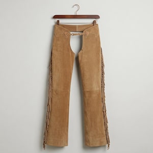 May include: Tan suede chaps featuring long fringe detailing along the outer legs, a high-waisted design with a central metal buckle closure at the waist, and a wide open-back construction, all suspended from a wooden clothes hanger.