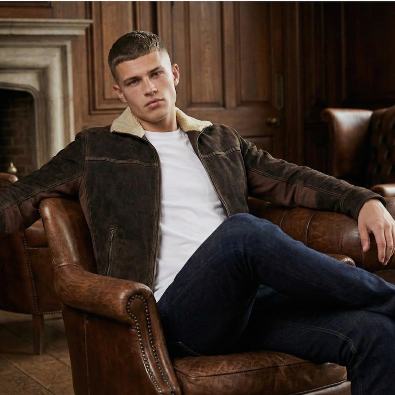 May include: A dark brown suede jacket with a cream-colored faux shearling collar, worn over a plain white t-shirt and dark blue denim jeans. The jacket features long sleeves, a front zipper closure, and visible stitching details along the chest.