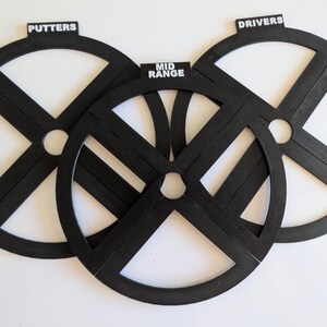 May include: Three black golf club guides with white centers are arranged on a white surface. The guides are labeled "PUTTERS", "MID RANGE", and "DRIVERS". The guides are circular with an "X" design.