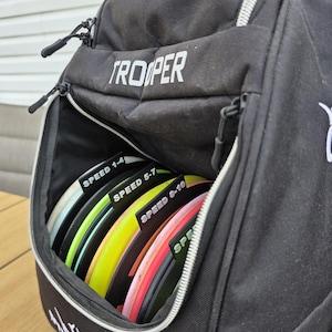 Puede incluir: Bolsa de golf de disco Trooper negra con un bolsillo frontal con cremallera lleno de discos coloridos. Los discos est&aacute;n etiquetados con rangos de velocidad: 1-4, 5-7 y 8-10. La bolsa est&aacute; sobre una superficie de madera.