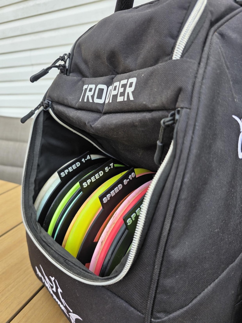 Puede incluir: Bolsa de golf de disco Trooper negra con un bolsillo frontal con cremallera lleno de discos coloridos. Los discos est&aacute;n etiquetados con rangos de velocidad: 1-4, 5-7 y 8-10. La bolsa est&aacute; sobre una superficie de madera.