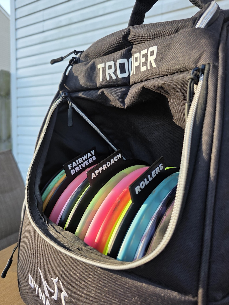 May include: Black "Trooper" disc golf bag with a zippered pocket filled with colorful discs. The discs are labeled "Fairway Drivers", "Approach", and "Rollers". The bag is open, revealing the discs inside. The bag has a handle and a zipper.