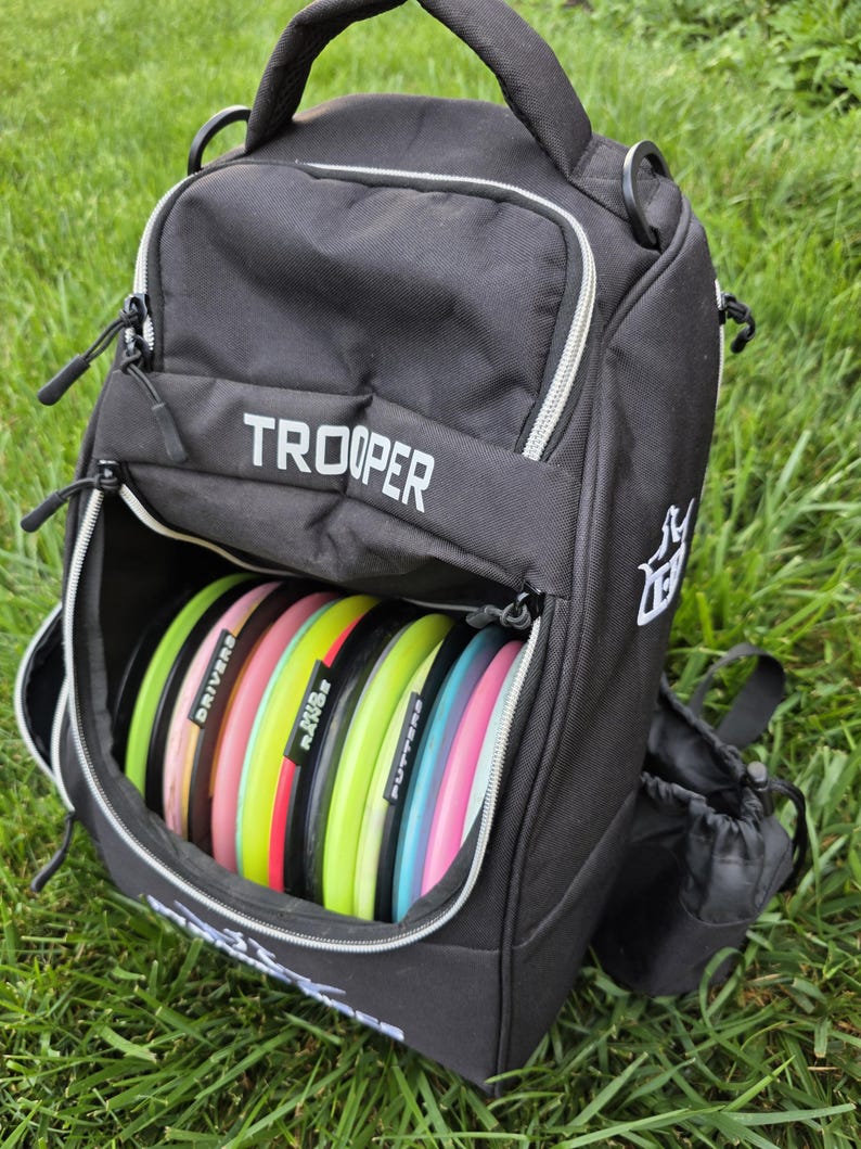 May include: Black disc golf bag with the word "TROOPER" in white. The bag is open, revealing a stack of colorful disc golf discs. The discs are labeled "DRIVERS", "RANGE", and "PUTTERS". The bag has a handle and a side pocket.