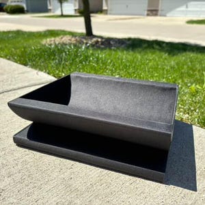 May include: A black, rectangular planter with a curved inner lip, sitting on a black, rectangular base. The planter is made of a textured material and appears to be made of concrete or a similar material.