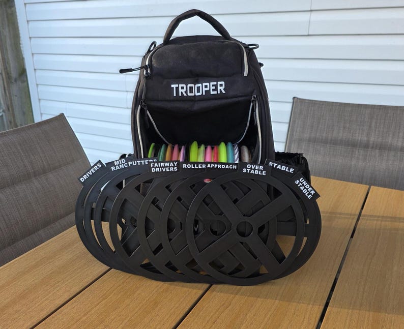 May include: A black disc golf bag labeled "TROOPER" is open, revealing colorful discs. In front are black disc golf flight path markers. The markers are labeled "DRIVERS", "MID RANGE PUTTERS", "FAIRWAY DRIVERS", "ROLLER APPROACH", "OVER STABLE", and "UNDER STABLE".