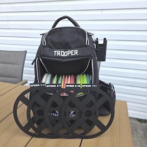 Puede incluir: Mochila negra de disc golf "Trooper" con una variedad de discos coloridos en el interior. Cuatro soportes de diana de disc golf negros est&aacute;n frente a la bolsa. La bolsa tiene un asa y un bolsillo lateral. El texto "SPEED 1-4", "SPEED 5-7", "SPEED 8-10" y "SPEED 11+" es visible.