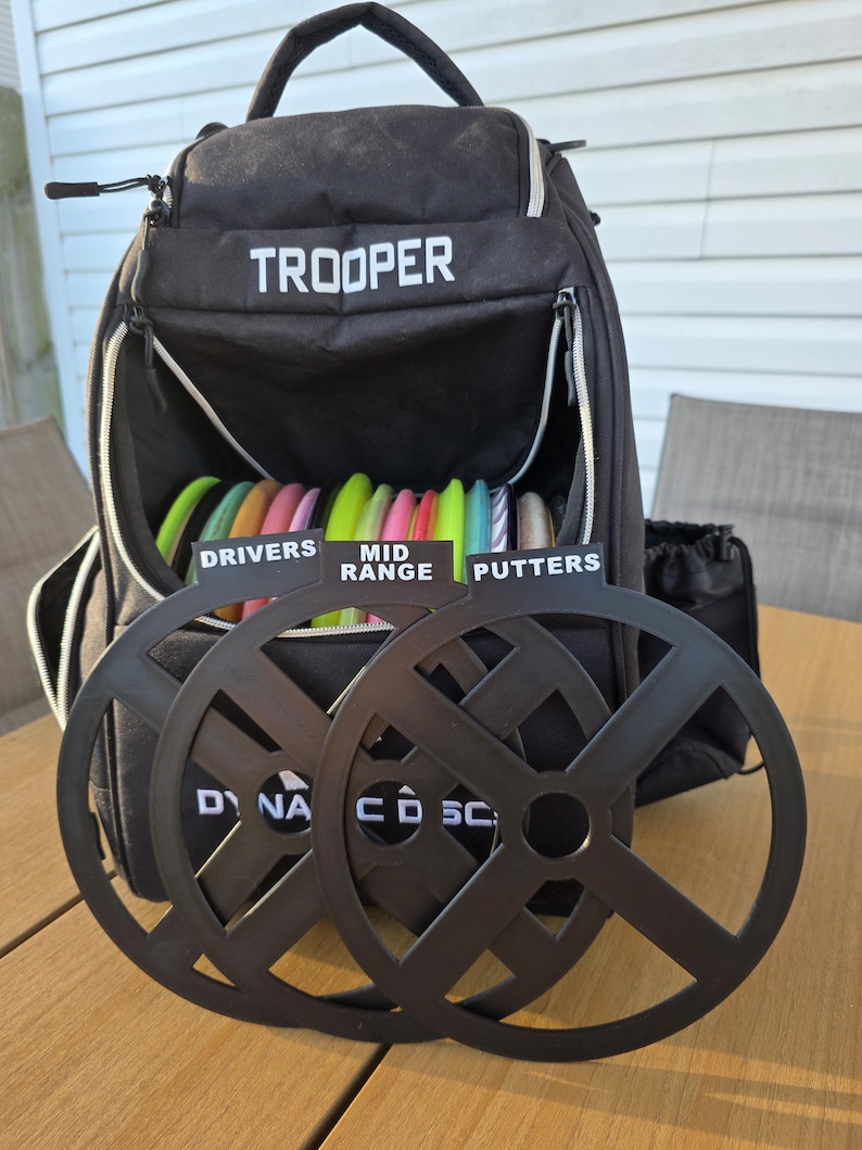 May include: Black disc golf backpack labeled "TROOPER" with a variety of colorful discs inside. Two black disc golf target baskets are in front of the backpack. The discs are labeled "DRIVERS", "MID RANGE", and "PUTTERS".