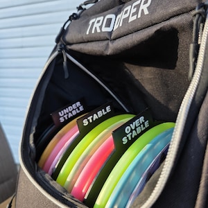 May include: Black "Trooper" disc golf bag with a zippered compartment holding several colorful disc golf discs. The discs are labeled "Under Stable", "Stable", and "Over Stable".