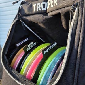 May include: Black disc golf bag with the word "TROOPER" in white. The bag is open, revealing colorful discs labeled "DRIVERS", "MID RANGE", and "PUTTERS". The discs are various colors including black, pink, yellow, and blue.