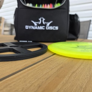 Puede incluir: Una bolsa negra de Dynamic Discs con logotipo blanco, un soporte de disco negro y un disco amarillo sobre una superficie de madera. La bolsa est&aacute; abierta, revelando discos coloridos en el interior. La escena sugiere una configuraci&oacute;n de disc golf.