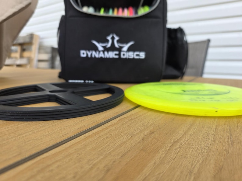 Puede incluir: Una bolsa negra de Dynamic Discs con logotipo blanco, un soporte de disco negro y un disco amarillo sobre una superficie de madera. La bolsa est&aacute; abierta, revelando discos coloridos en el interior. La escena sugiere una configuraci&oacute;n de disc golf.