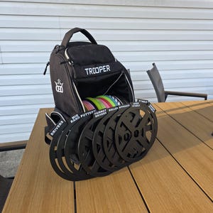 May include: Black Trooper disc golf bag with a white logo and the word "TROOPER" in white. The bag is open, revealing colorful discs. Black disc holders are in front of the bag, labeled "DRIVERS", "MID RANGE PUTTERS", "FAIRWAY DRIVERS", "ROLLER APPROACH", and "OVER STABLE".