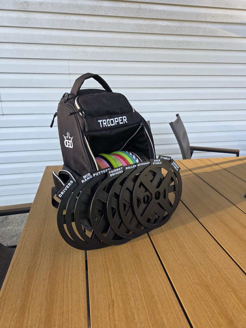 May include: Black Trooper disc golf bag with a white logo and the word "TROOPER" in white. The bag is open, revealing colorful discs. Black disc holders are in front of the bag, labeled "DRIVERS", "MID RANGE PUTTERS", "FAIRWAY DRIVERS", "ROLLER APPROACH", and "OVER STABLE".