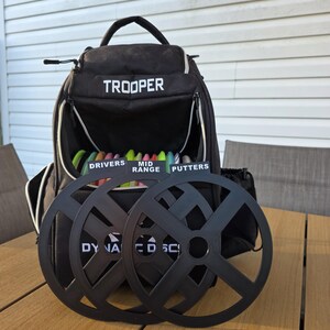 May include: Black "Trooper" backpack with white trim, open to reveal colorful disc golf discs. Two black disc golf disc wheels are in front of the bag. The bag has the words "Drivers", "Mid Range", and "Putters" on the inside.