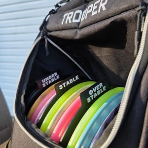 May include: Black disc golf bag with the word "TROOPER" in white. The bag is open, revealing a stack of colorful disc golf discs. Each disc is labeled with "UNDER STABLE", "STABLE", and "OVER STABLE".