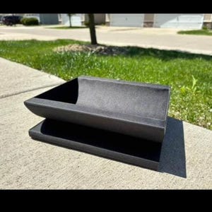 May include: A dark gray, rectangular planter box with a curved top section, resting on a flat base. The planter is made of a textured material and sits on a concrete surface, with green grass and a residential background.