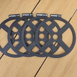 Puede incluir: Cuatro objetos circulares negros con un dise&ntilde;o en forma de X, cada uno etiquetado con un rango de velocidad: 1-4, 5-7, 8-10 y 11+. Los objetos est&aacute;n dispuestos sobre una superficie de madera marr&oacute;n claro.