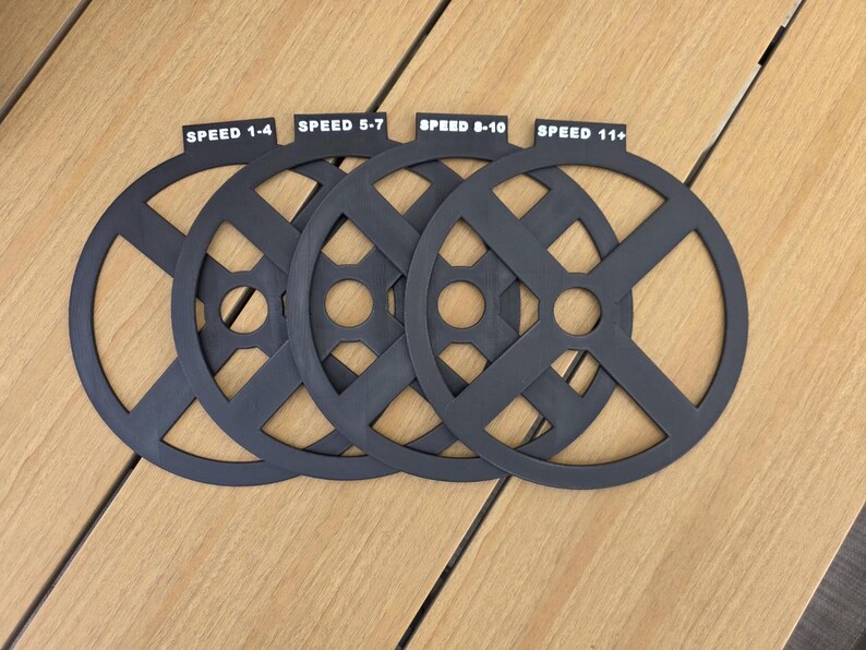 Puede incluir: Cuatro objetos circulares negros con un dise&ntilde;o en forma de X, cada uno etiquetado con un rango de velocidad: 1-4, 5-7, 8-10 y 11+. Los objetos est&aacute;n dispuestos sobre una superficie de madera marr&oacute;n claro.