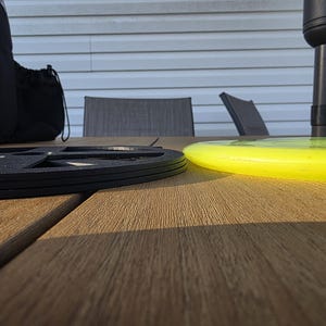 May include: A black disc golf basket base and a bright yellow disc are on a wooden table. A black disc golf bag is in the background. The scene is outdoors, with a white siding and a patio umbrella visible.