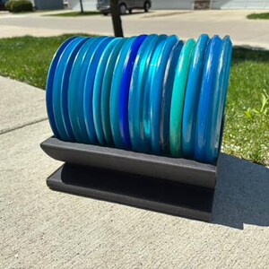 May include: A stack of blue and green disc golf discs on a black stand. The discs are arranged in a gradient from dark blue to light green.