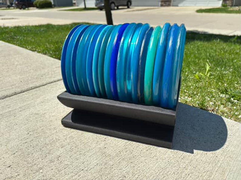 May include: A stack of blue and green disc golf discs on a black stand. The discs are arranged in a gradient from dark blue to light green.