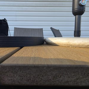 May include: A stack of black coasters sits on a brown wooden table, next to a light-colored, weathered wooden object. The scene is outdoors, with a partial view of a black umbrella and a gray chair in the background.