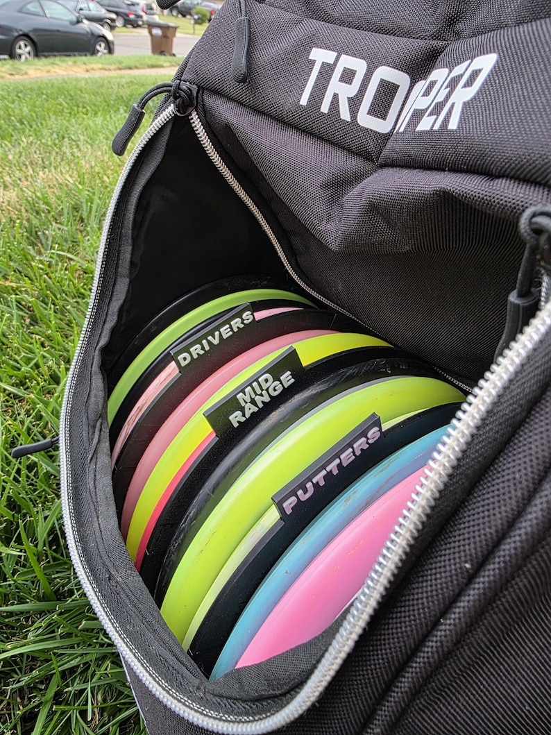 May include: Black disc golf bag with the word "TROOPER" in white. The bag is open, revealing a stack of colorful disc golf discs. The discs are labeled "DRIVERS", "MID RANGE", and "PUTTERS".