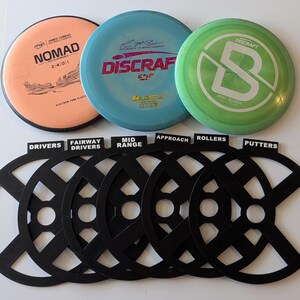 May include: Six black disc golf disc organizers with labels for drivers, fairway drivers, mid-range, approach, rollers, and putters. Three disc golf discs are visible in the background: a pink disc with "Nomad" written on it, a blue disc with "Discraft" written on it, and a green disc with a white "B" on it.