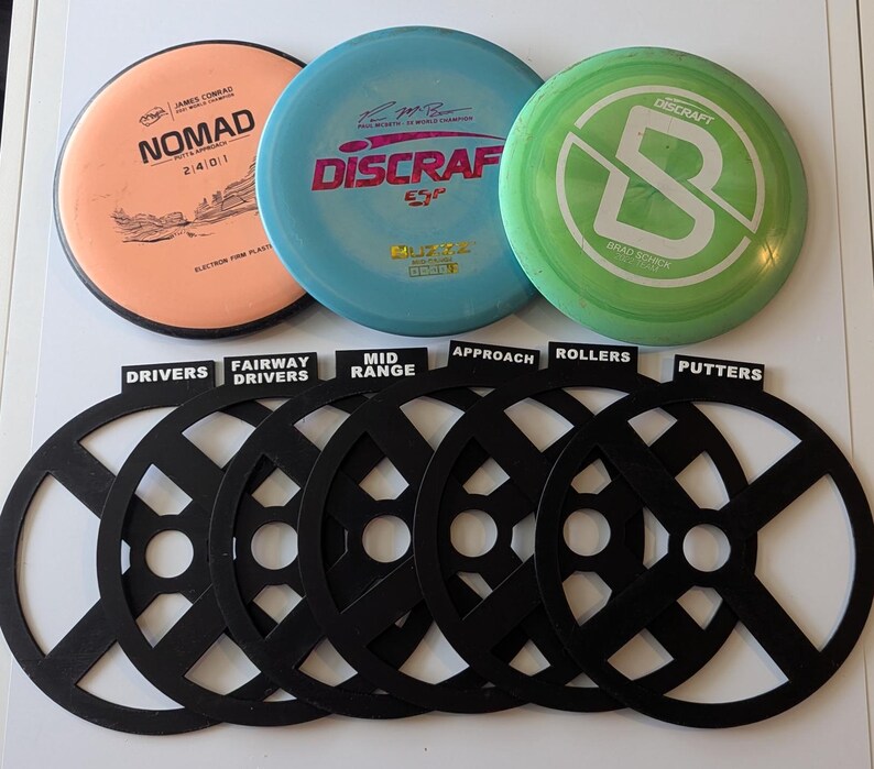 May include: Six black disc golf disc organizers with labels for drivers, fairway drivers, mid-range, approach, rollers, and putters. Three disc golf discs are visible in the background: a pink disc with "Nomad" written on it, a blue disc with "Discraft" written on it, and a green disc with a white "B" on it.