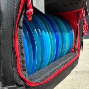 May include: A black disc golf bag with a red zipper and a compartment for storing discs. The compartment is filled with blue and green discs.