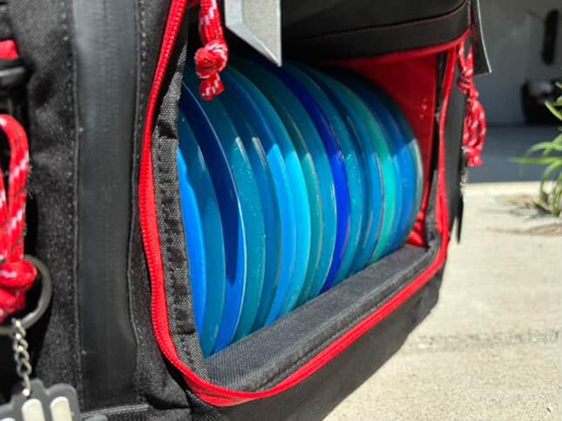 May include: A black disc golf bag with a red zipper and a compartment for storing discs. The compartment is filled with blue and green discs.
