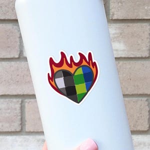 May include: White water bottle with a black lid and handle. A sticker of a heart-shaped design with a checkered pattern in black, green, and blue, surrounded by flames in red, orange, and yellow. The bottle is held against a brick wall.