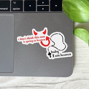May include: A laptop with three stickers. One sticker has the text "i don't think this nun is going to heaven" with a devil graphic. Another sticker has a rosary and the text "baby i am heaven". The third sticker is a white shape.