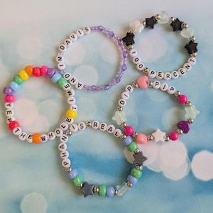 May include: Five circular beaded bracelets featuring colorful plastic beads, letter beads spelling phrases like YEP THE WHOLE TIME IS REAL, AND IM DAVIS, 100 PERCENT, and PHAN IS REAL, along with star and heart-shaped charms in various colors.