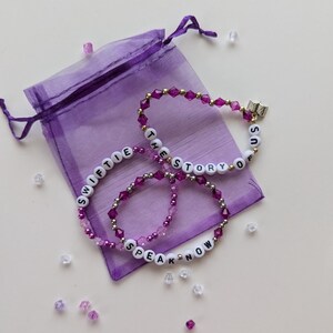 Several Taylor Swift friendship bracelets on a purple organza gift bag on a white background. The bracelets have lyrics and titles from Taylor Swift's album Speak Now including The Story of Us.