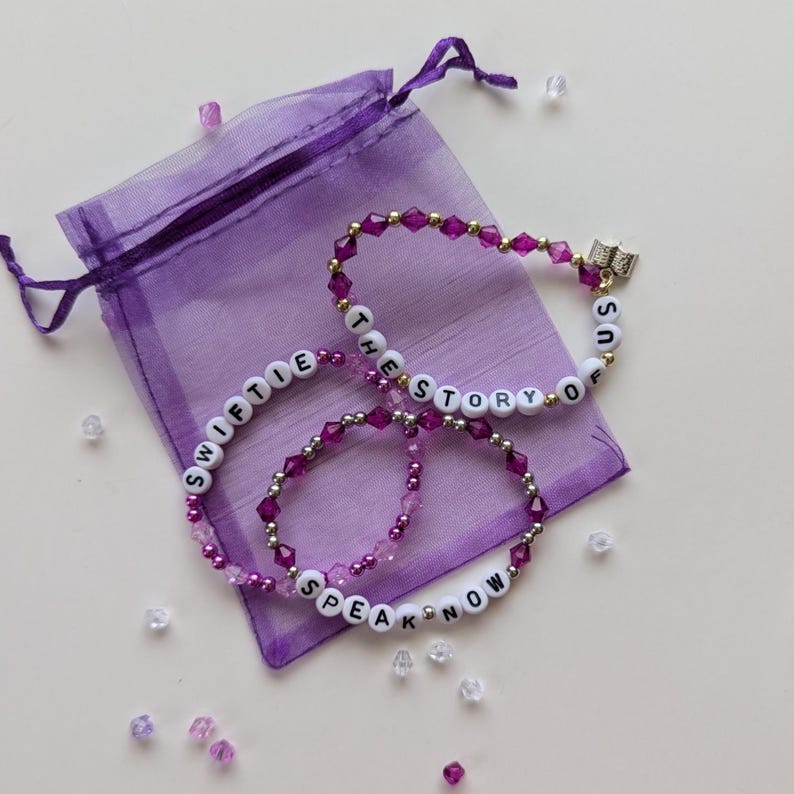Several Taylor Swift friendship bracelets on a purple organza gift bag on a white background. The bracelets have lyrics and titles from Taylor Swift's album Speak Now including The Story of Us.