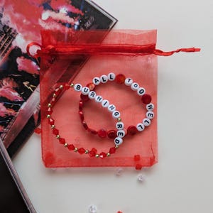 Several Taylor Swift friendship bracelets on a red organza gift bag on a white background. The bracelets have lyrics and titles from Taylor Swift's album Red including All Too Well and Burning Red.