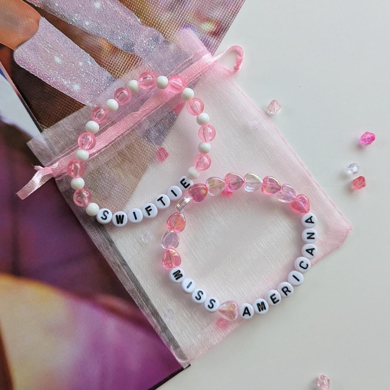 Several Taylor Swift friendship bracelets on a pink organza gift bag on a white background. The bracelets have lyrics and titles from Taylor Swift's album Lover including Miss Americana.